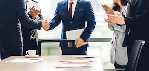 Image two business partners in suits shaking together in front of group of people clapping hands in modern office.